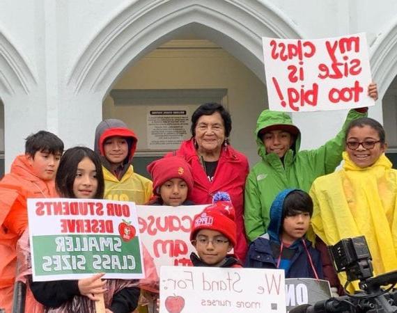 学生们和Dolores Huerta站在一起，穿着雨衣，举着红色的爱德标志